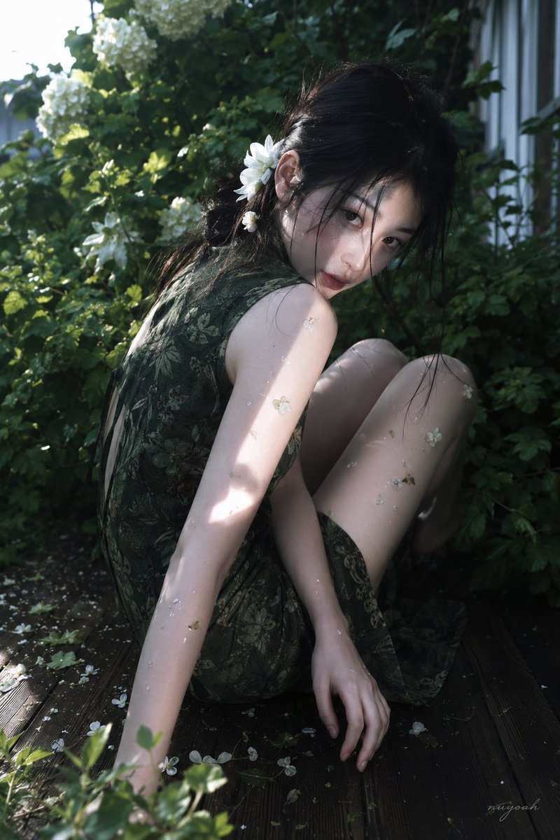 真人摄影,真实人物,户外花园唯美人像写真,低饱和冷绿调与斑驳自然光,轻微胶片柔焦和电影感。竖幅近中景,略高机位俯拍,人物占画面约70%,身体斜向贯穿画面,前景为木质露台和少量植物虚化,背景是深绿色灌木、白色绣球花与隐约建筑边缘。年轻亚洲女性,黑色长发低束且略凌乱,脸侧有细碎发丝,发间和耳后插白色花朵;清秀柔和五官,冷白到象牙白肤调,面颊与唇色带淡粉血色,阴影处受绿植反光轻微偏青。身形纤细修长,肩颈轻薄,手臂线条细长,腿部弯曲形成柔和曲线。她侧坐蜷曲在木平台上,一条腿抬起弯曲,身体前收,左臂向前撑地,另一只手自然贴在身侧木板上,头部低垂并回望镜头;眼神湿润安静,眉眼微垂,嘴唇轻闭,表情克制、羞怯、带一点疲惫疏离。穿深绿色无袖改良中式短裙,布料薄而柔软,暗纹植物纹理,哑光微反射,贴合上身与腰部,坐姿处产生自然拉伸、细小褶皱和裙摆堆叠。皮肤、手臂和腿部散落少量白色花瓣与碎屑,阳光从左上方穿过树叶形成硬边光斑,照亮肩颈、手臂和腿部,暗部柔和偏冷,背景浅景深虚化,真实光影、细腻皮肤高光、轻微颗粒与压缩感