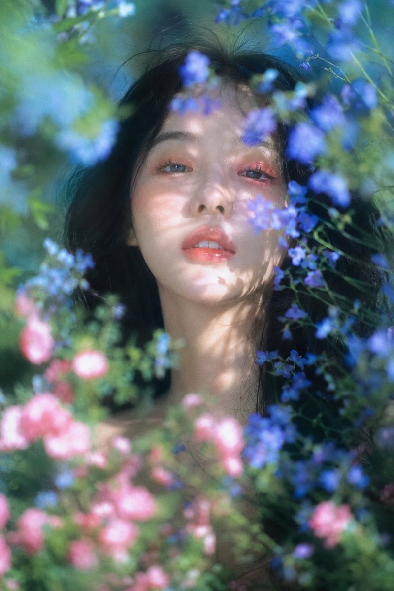 梦幻自然光花园人像,柔焦胶片质感,竖幅近距离肖像构图;年轻女性,muse般的面庞,黑色长发蓬松微乱,面部居中偏上作为主视觉,白皙偏暖象牙肤调,橘粉眼妆与水润珊瑚唇,正面安静凝视镜头,头部略抬,嘴唇轻启,眼神湿润疏离、无笑意;身体只露出少量肩颈,被花枝遮挡。前景粉色、蓝紫色小花和绿色枝叶中等密度环绕面部并局部遮挡,背景蓝绿色自然虚化。上方斜侧日光穿过花枝,在脸颊、鼻翼、下巴 and 颈部形成真实斑驳叶影,皮肤细腻柔亮,鼻梁和唇部有自然高光,浅景深,前景花朵虚化,轻微胶片颗粒
