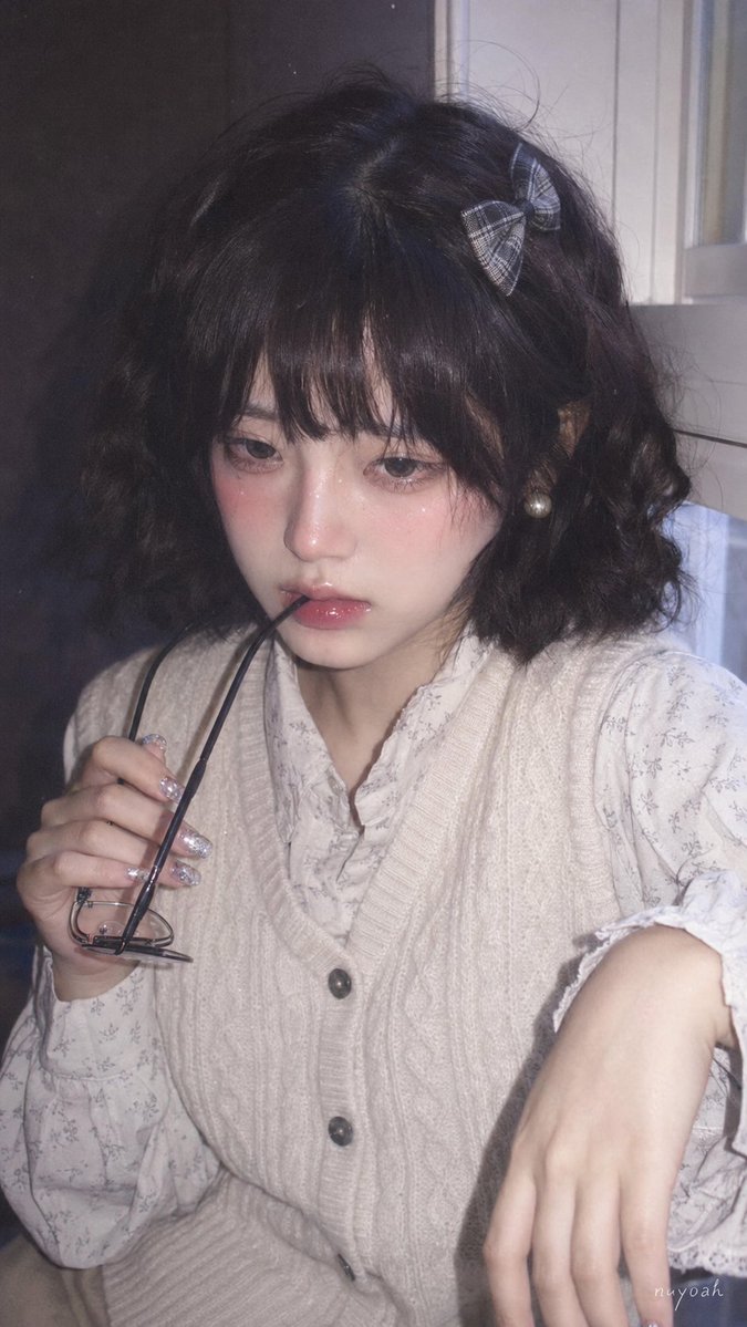 真人摄影、真实人物、日系复古 Pin 图气质的柔焦手机闪光人像,竖幅 9:16,近景半身构图,略高于眼平的正面机位,人物居中偏右,脸部占据上半部主视觉,右侧白色窗框作为辅助竖向结构,左侧深色室内墙面压暗背景。年轻女性,冷白到象牙白肤调,脸颊和鼻尖有粉红腮红与细小闪粉,眼神湿润安静、略带羞怯和无辜感,眉眼放松,嘴唇微张并含住黑色细框眼镜的一端,嘴角不笑。蓬松黑棕短卷发、空气感齐刘海,右侧格纹蝴蝶结发夹,珍珠耳钉,亮泽唇釉,银色闪片美甲;一只手抬起眼镜靠近唇边,另一只手搭在右侧前景,身体轻微前倾、肩颈前收。穿米白粗针织马甲叠搭浅色印花衬衫,马甲罗纹和纽扣边缘自然贴合,上身有柔和包裹感,衬衫领口袖口产生轻薄褶皱,针织哑光绒感与衬衫低反光材质清晰。右侧窗光混合正面闪光,冷白高光、灰紫暗部、低饱和粉米色调,局部过曝和朦胧光晕,保留脸部水润高光、唇部反光、指甲闪片反射;CCD/手机压缩质感,轻微噪点、灰尘颗粒、柔焦低对比,不要动漫、不要插画