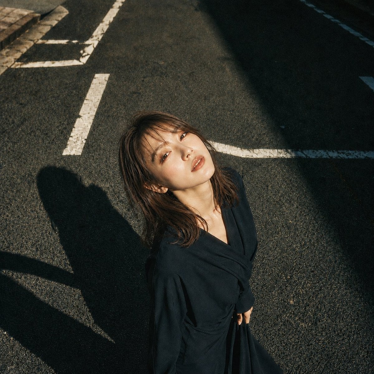 主題:白線と影 人物・表情: 20代女性。明るいアイボリーベージュ寄りの白肌に、ほんのりピンクの血色を含んだ自然な肌質。頬、鼻先、鎖骨に柔らかなハイライト。精密で上品なドールライク日系メイク、淡いローズピンクのチーク、シャンパンベージュとピンクブラウンの目元、細く長いまつ毛、自然な涙袋、明るめブラウンの柔らかい眉、透け感のあるピンクベージュのグロスリップ。瞳に小さなキャッチライト。静かに見上げる、少し余韻のある表情。 服装・ポーズ: 深い黒に近いネイビーの和モダンなラップワンピース。長袖、重なりのあるVライン、腰で布を結んだようなドレープ、落ち感のある柔らかな生地。身体は画面下側に立ち、上半身を少し後ろへ預ける。顔はカメラへ向けて上げ、右手はスカートの布を軽く持ち、左腕は自然に体側へ落とす。姿勢は大人っぽく、静かな緊張感を残す。 背景・光: 日本の都市のアスファルト道路。白い道路標示が斜めに入り、画面左上に歩道の縁石が少し見える。強い午後の太陽が画面左上から差し、顔と髪、胸元を明るく照らす。人物の影は画面左下へ長く伸び、画面右上には建物の濃い影が大きく落ちる。白線、影、アスファルトの粒感をはっきり描写する。 構図・カメラ: 1:1の正方形。高い位置から見下ろす俯瞰ポートレート。人物は画面下中央寄りに配置し、道路の余白を広く残す。カメラは真上より少し斜め、人物の視線はレンズへ向かう。中距離のストリートポートレート、背景まで質感が残る自然な被写界深度。…