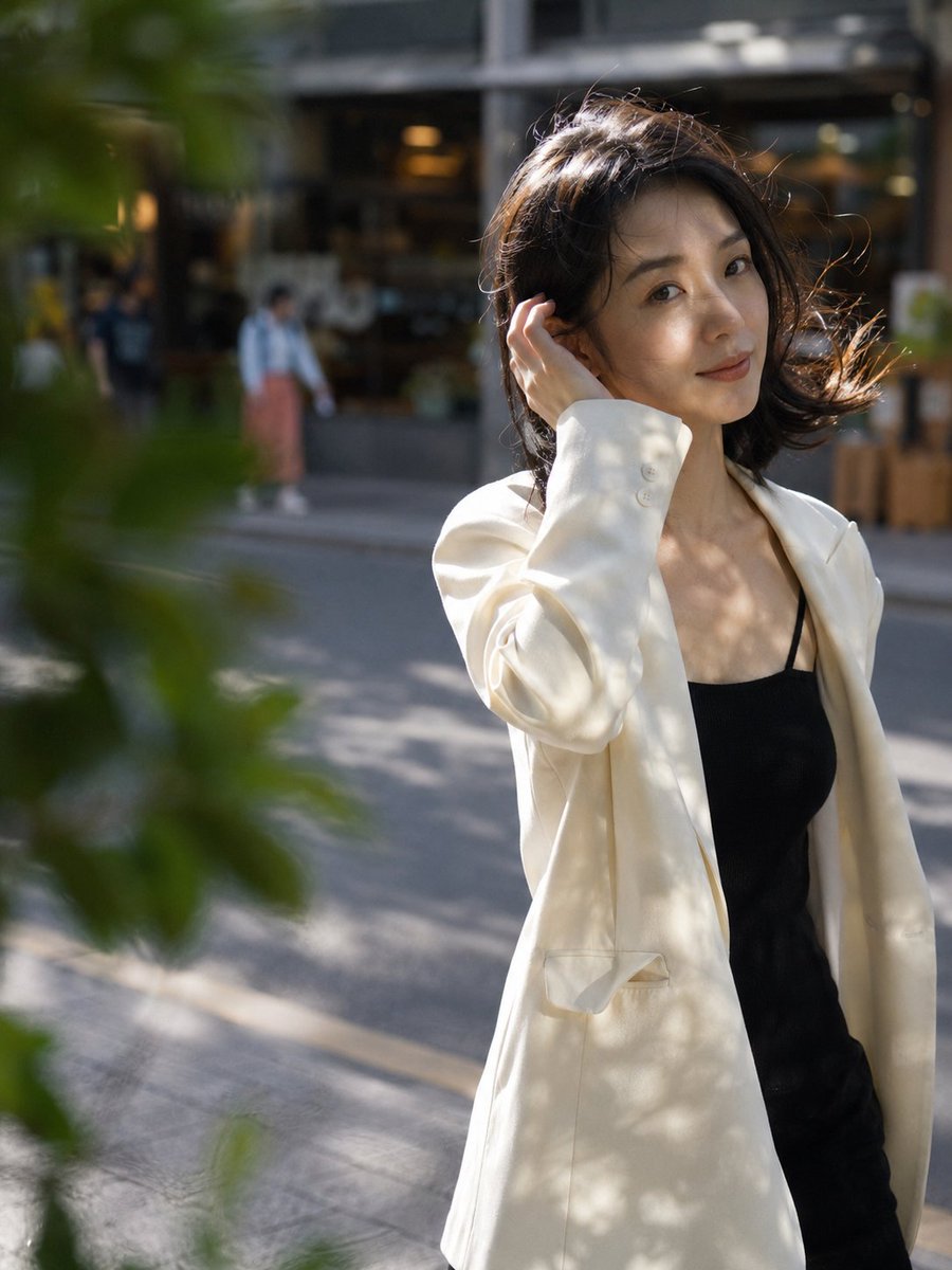 主体:一位25岁左右的中国女性,鹅蛋脸,黑色及肩微卷长发被风吹起几缕,穿米白色oversized西装外套内搭黑色吊带,锁骨清晰 动作与姿态:走在人行道上被镜头抓拍的瞬间,一只手轻拨耳边碎发,身体微微侧转,步伐自然 表情与视线:不经意间瞥向镜头的瞬间,嘴角似笑非笑,眼神松弛真实 场景环境:下午四点的城市街头,梧桐树影落在人行道上,背景是虚化的咖啡店橱窗和路人 镜头:中景(medium shot),85mm镜头,浅景深(shallow DOF),主体位于画面右三分之一,前景有虚化的树叶 光线:午后侧逆光(backlight)勾勒发丝金边,树影在脸上形成斑驳光斑(dappled light),自然柔和…