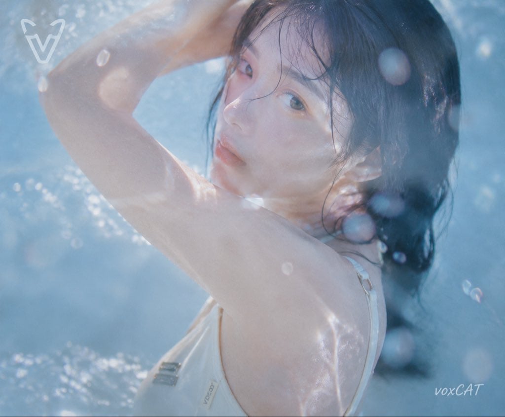日系夏日水感写真,一位年轻女性角色,清透少女气质,湿润黑色长发,自然妆容,穿浅色简约泳装。 采用隔水拍摄视角,镜头位于水面或透明介质后方,前景出现水波折射、水雾、玻璃反光遮挡,人物被包裹在透明蓝色空间中。 人物半身动态构图,手臂抬起形成自然弧线,身体轻微转动,湿发贴脸,像夏日电影中的瞬间抓拍。 泳池蓝色环境,下午自然光,冷蓝水光包围人物,暖肤色,高曝光,低饱和,柔焦,朦胧梦幻质感,日系青春电影截图,老DV录像感,水下摄影效果。 整体加入VOXCAT品牌元素: 左上角加入简洁现代图形logo,由stylized“V”和猫元素结合构成,白色半透明质感,与水雾融合; 右下角加入小型签名“voxCAT”,vox小写、CAT大写; 服装局部加入低调VOXCAT织标、金属铭牌或暗纹压印。 梦幻夏日氛围,透明感摄影,胶片质感。…